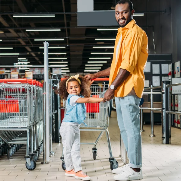 近くのスーパー マーケットのトロリーのショッピングの娘と一緒に立っているアフリカ系アメリカ人の笑顔  — 無料ストックフォト