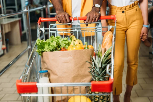 スーパーで食品のショッピング トロリーを運ぶ若いカップルの画像をトリミング — ストック写真