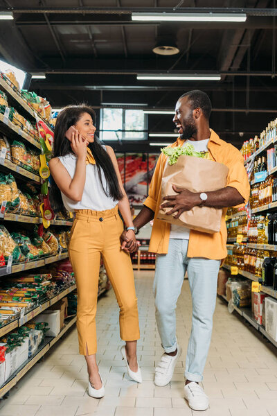 laughing african american man with paper bag holding hand of girlfriend while she talking on smartphone in supermarket