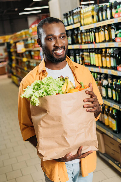 smiling african american male shopper holding paper bag with fruits and vegetables in supermarket