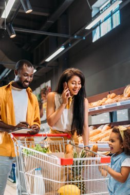 gülümseyen Afrikalı-Amerikalı kadın arabası süpermarkette alışveriş ile yakın duran kızı ve eşi ise Smartphone cep telefonu ile konuşmak 