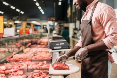 cropped image of african american male shop assistant in apron cutting raw meat in grocery store  clipart