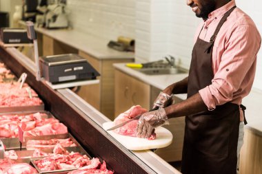 partial view of smiling african american male shop assistant in apron cutting steak of raw meat in hypermarket clipart