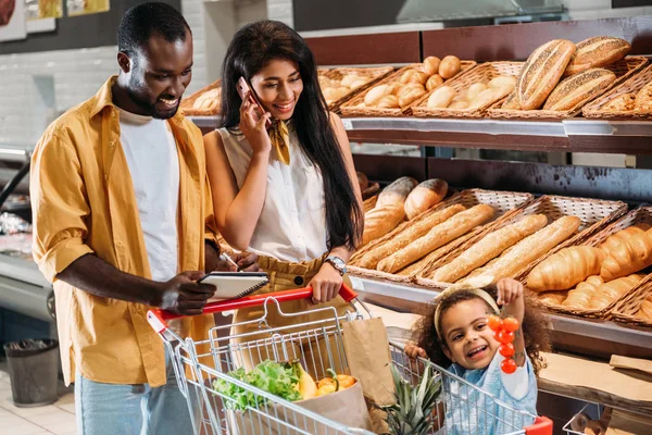 非裔美国妇女谈论智能手机 而她的丈夫和女儿站在附近与购物车在超级市场 — 图库照片