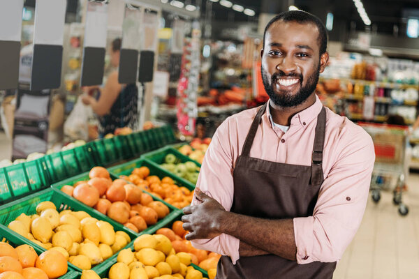 african american male shop assistant in apron standing with crossed arms near fruit department in hypermarket 
