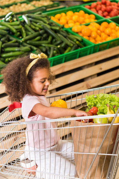 happy african american child sitting in shopping trolley in grocery store 