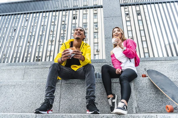 若い街でスケート ボードの近くのハンバーガーを食べる文化のカップルを笑顔の低角度のビュー — ストック写真