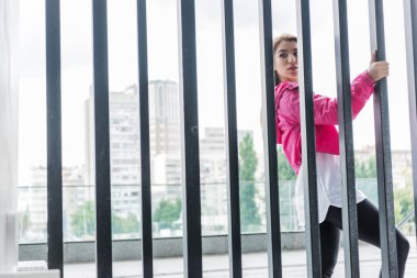 attractive stylish asian woman standing near metal fence and looking away at urban street clipart