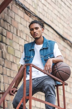 stylish mixed race man standing with basketball ball and looking at camera at street clipart
