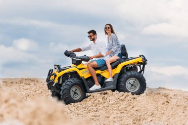 happy young couple riding all-terrain vehicle in desert on cloudy day clipart