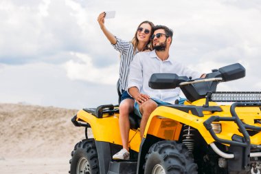 attractive young couple taking selfie while sitting on ATV in desert clipart