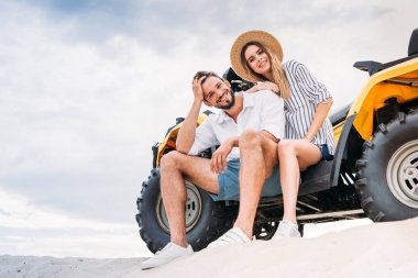 beautiful young couple sitting on ATV on sandy dune and looking at camera clipart