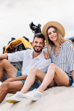 mutlu genç çift oturma Atv kabuklarıyla kumlu yakınındaki ve kameraya bakıyor