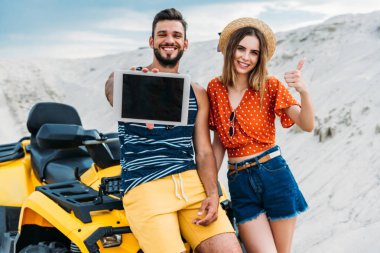 smiling young couple with atv showing digital tablet with blank screen and thumb up at desert clipart