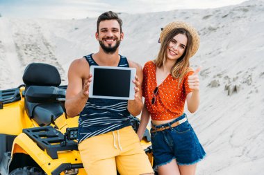 beautiful young couple with atv showing digital tablet with blank screen and thumb up at desert clipart