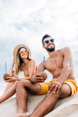 bottom view of young couple with coconut cocktails sitting on sand in front of cloudy sky clipart