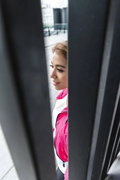 都市のストリートで金属フェンス近くに立っている笑顔のアジア女性の選択と集中  — 無料ストックフォト