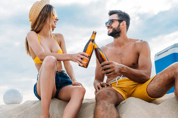beautiful young couple clinking with bottles of beer while sitting on sand