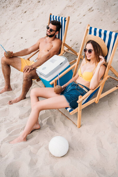 high angle view of beautiful young couple with coconut cocktails relaxing in sun loungers on sandy beach