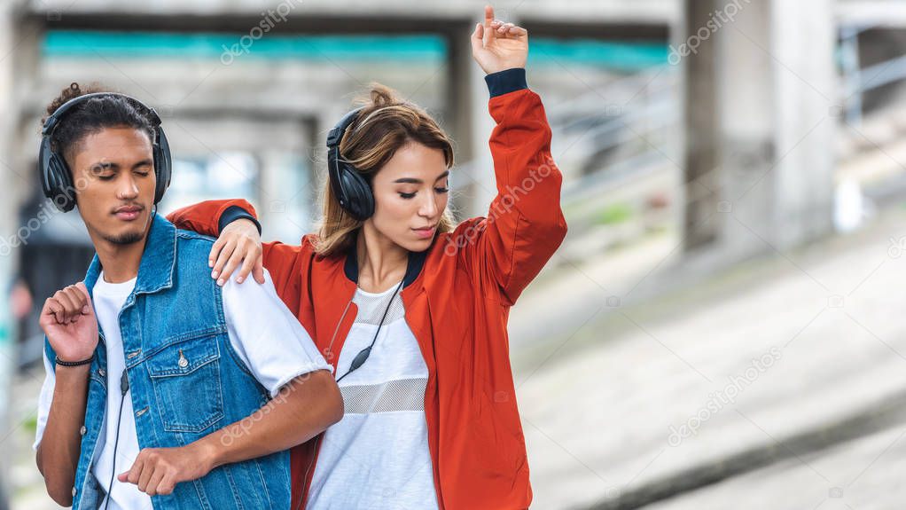 young multicultural couple listening music with headphones and dancing at city street 