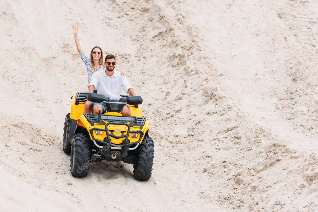 happy young couple riding ATV in desert and waving at camera
