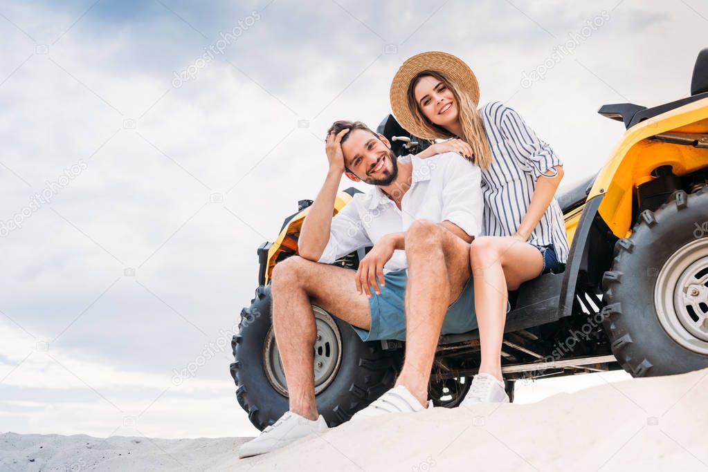 beautiful young couple sitting on ATV on sandy dune and looking at camera