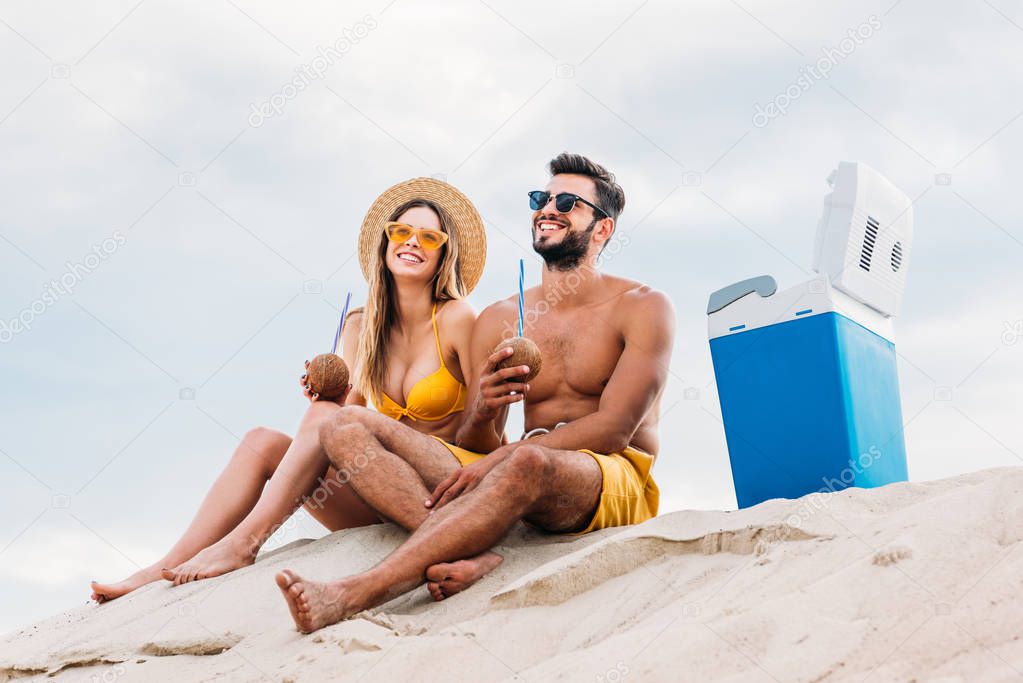 smiling young couple with coconut cocktails relaxing on sand in front of cloudy sky