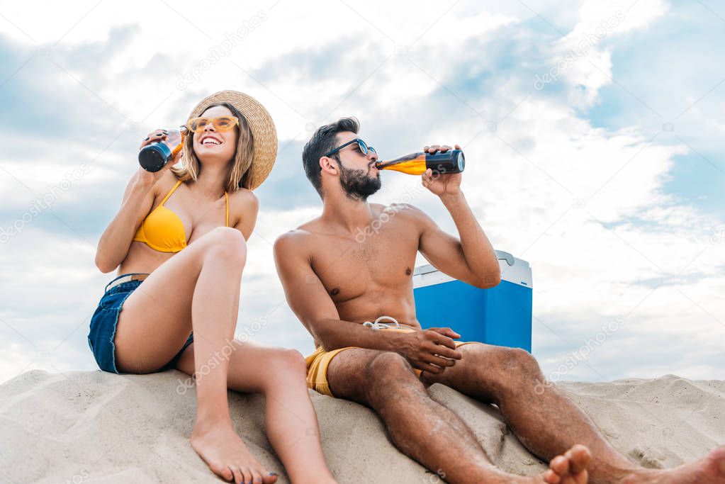 beautiful young couple drinking beer while sitting on sand