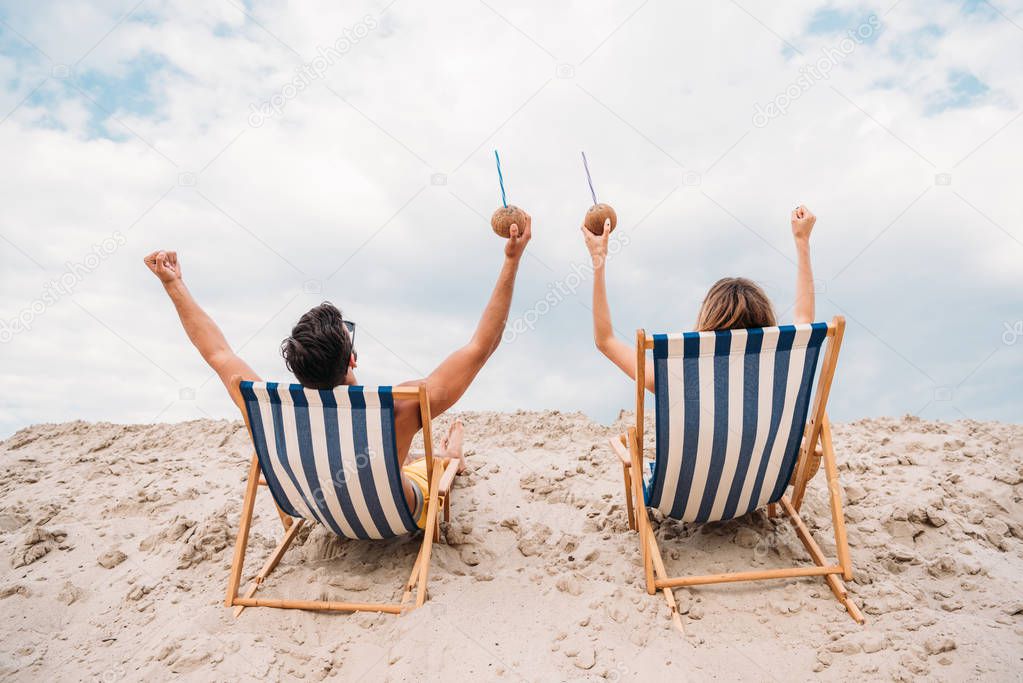 rear view of young couple with coconut cocktails relaxing in sun loungers on sandy dune