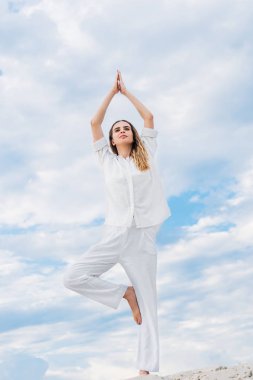 fit young woman practicing yoga in tree pose while standing on sand dune clipart