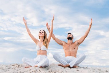 lotus poz (padmasana) yükseltilmiş kollu kabuklarıyla kumlu oturan güzel genç çift