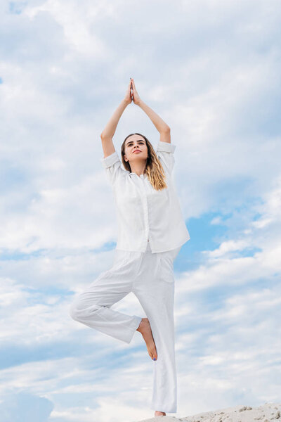 fit young woman practicing yoga in tree pose while standing on sand dune