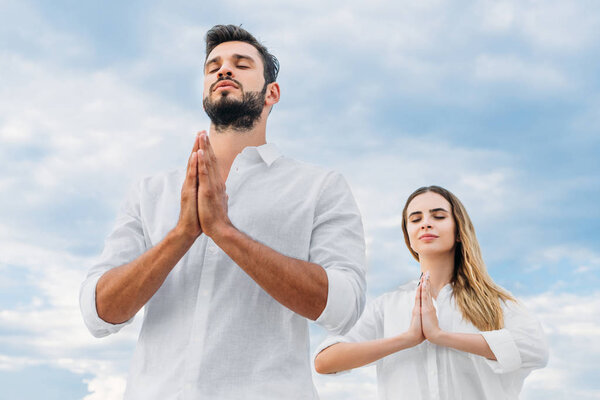bottom view of young couple meditating and making namaste mudra against cloudy sky
