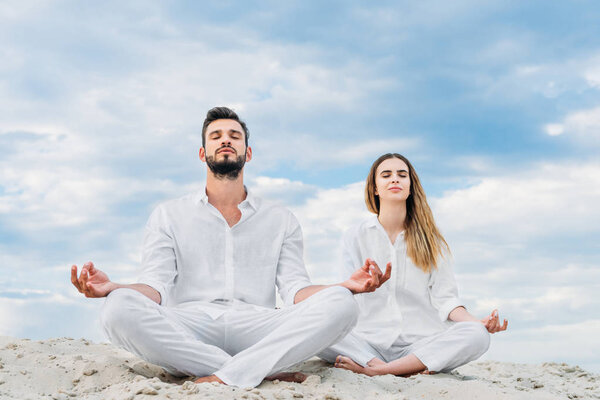 calm young couple meditating while sitting on sandy dune in lotus pose (padmasana)