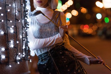 cropped shot of stylish young woman with golf club leaning on wall with white garland on city street at night clipart