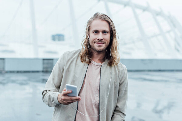 handsome young man listening music with earphones and smartphone on street and looking at camera