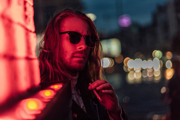 attractive young man in sunglasses smoking cigarette under red light on street