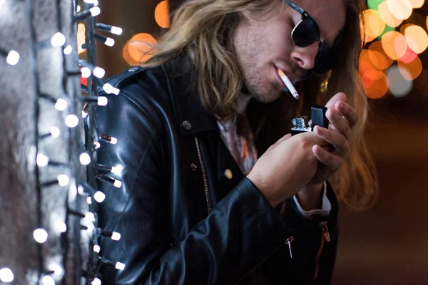 サングラスと革ジャケットは夜の通りにガーランドの下でタバコを吸ってでハンサムな若い男 — ストック写真