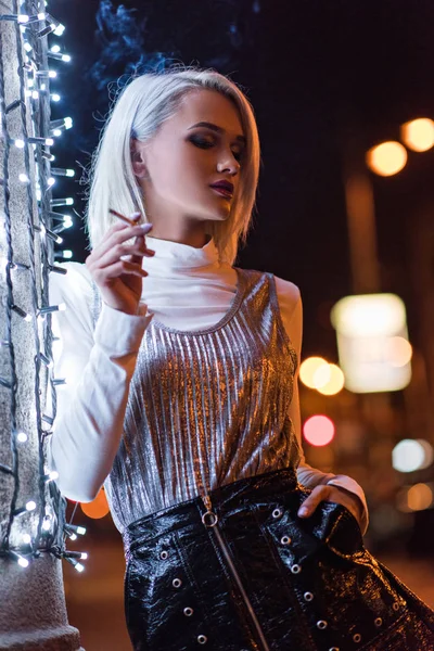 美しい若い女性白ガーランドで壁にもたれながら夜路上喫煙 — ストック写真