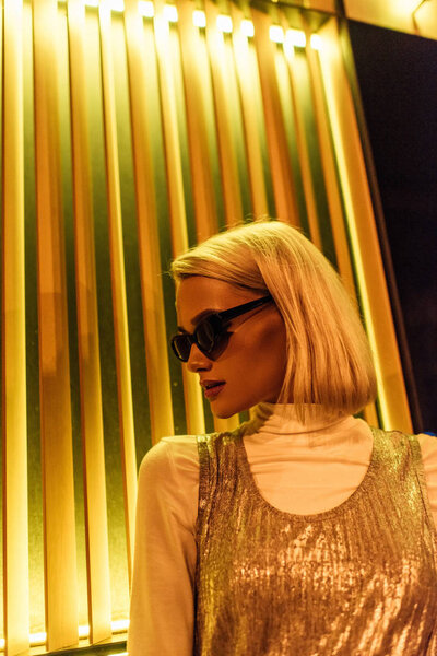 close-up portrait of stylish young woman in sunglasses and glossy top over turtleneck on street at night under yellow light