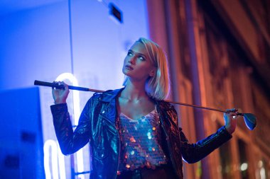 attractive young woman in leather jacket standing on street at night under blue light and holding golf club clipart