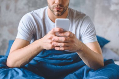 Genç adamın evde yatakta smartphone kullanarak resim kırpılmış