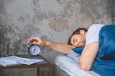 young sleeping man in bed turning off alarm clock at home  clipart
