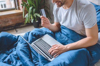 bir dizüstü bilgisayarda çevrimiçi alışveriş yaparken kredi kartıyla yatakta genç adam kırpılmış görüntü 