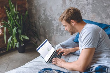 Genç adam yatakta dizüstü bilgisayar ekranında facebook ile kullanarak