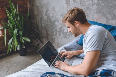 yakışıklı genç adam yatakta dizüstü ekran üzerinde rezervasyon web sitesi ile kullanma