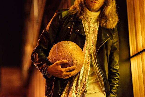 cropped shot of stylish man in leather jacket holding golden basketball ball under yellow light on street at night