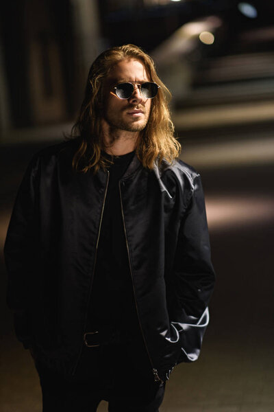 attractive young man in sunglasses and leather jacket on city street at night