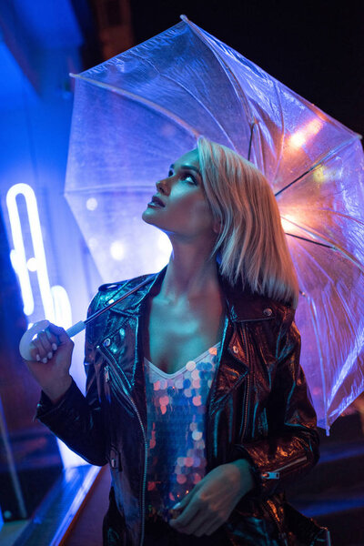 attractive young woman with umbrella looking up on street at night under blue light