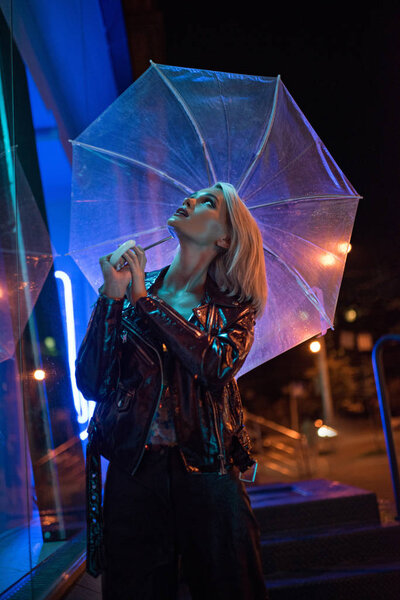 beautiful young woman with umbrella looking up on street at night under blue light